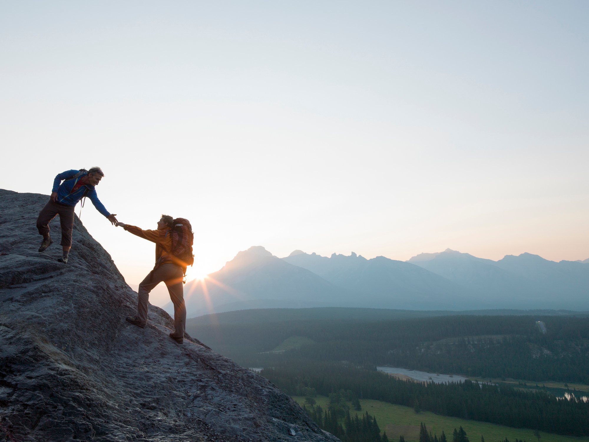 Two hikers at sunset, one reaching back to help another climb